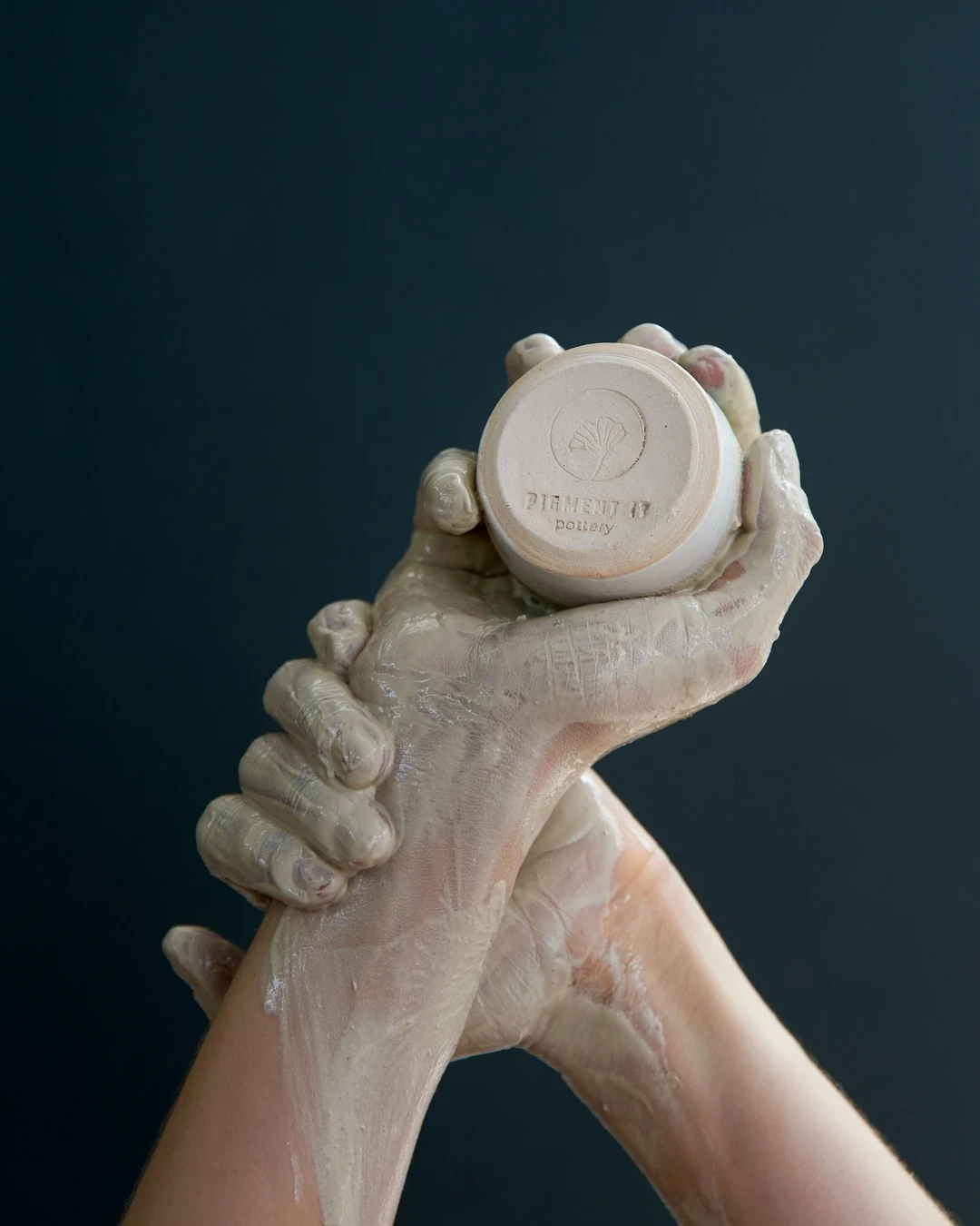 Mains couvertes d’argile tenant une poterie marquée du logo Pigment It Pottery.